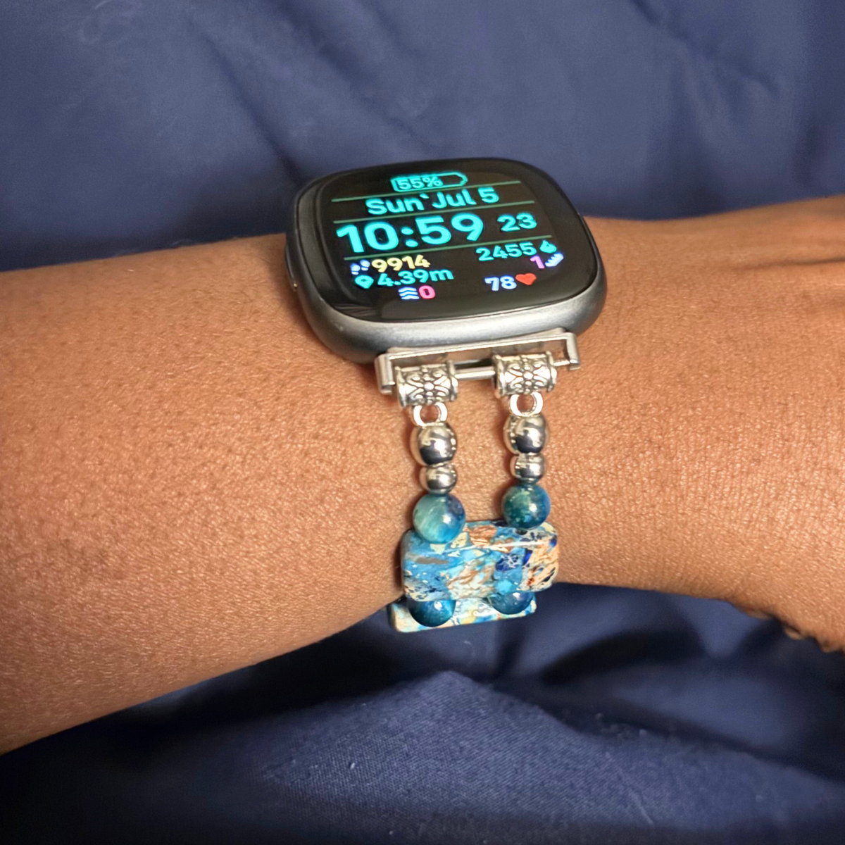A close-up of a smartwatch worn on a person's wrist, featuring a silver-toned charm bracelet strap with blue beads and a decorative blue-and-gold pendant, set against a dark blue fabric background.