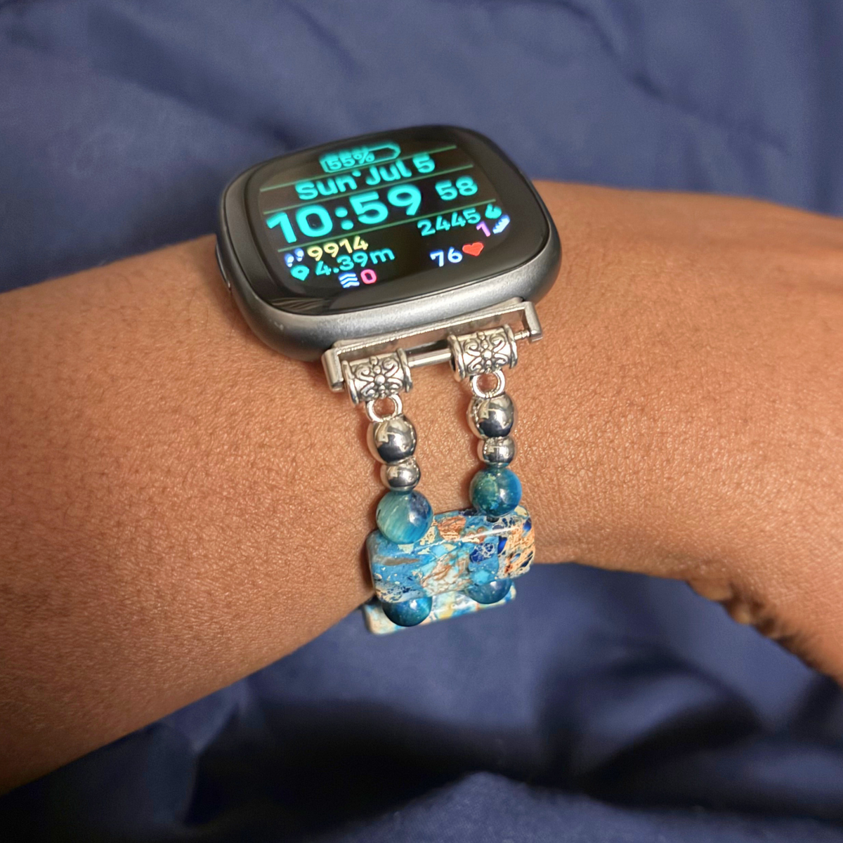 A close-up of a smartwatch worn on a person's wrist, featuring a silver-toned bracelet with blue marbled beads and a decorative blue and white patterned charm, set against a dark blue fabric background.