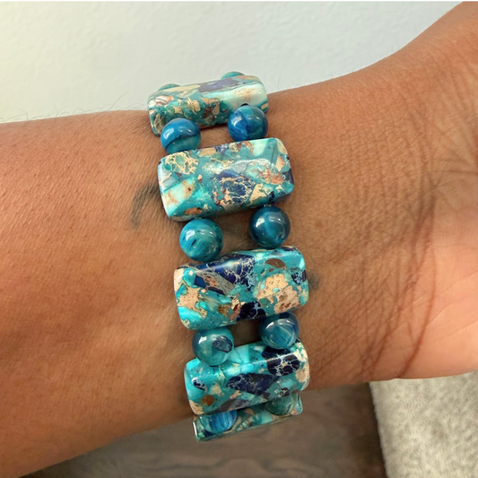 A close-up of a person's wrist wearing a stretch bracelet made of turquoise-blue marbled rectangular beads with small round blue beads spaced throughout.